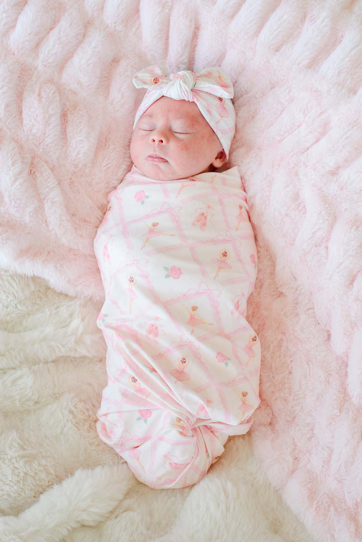 Sleeping newborn baby swaddled in a Caden Lane ballerina and roses pink print swaddle blanket with matching bow headwrap, laying next to a stuffed bunny on a pink fur blanket
