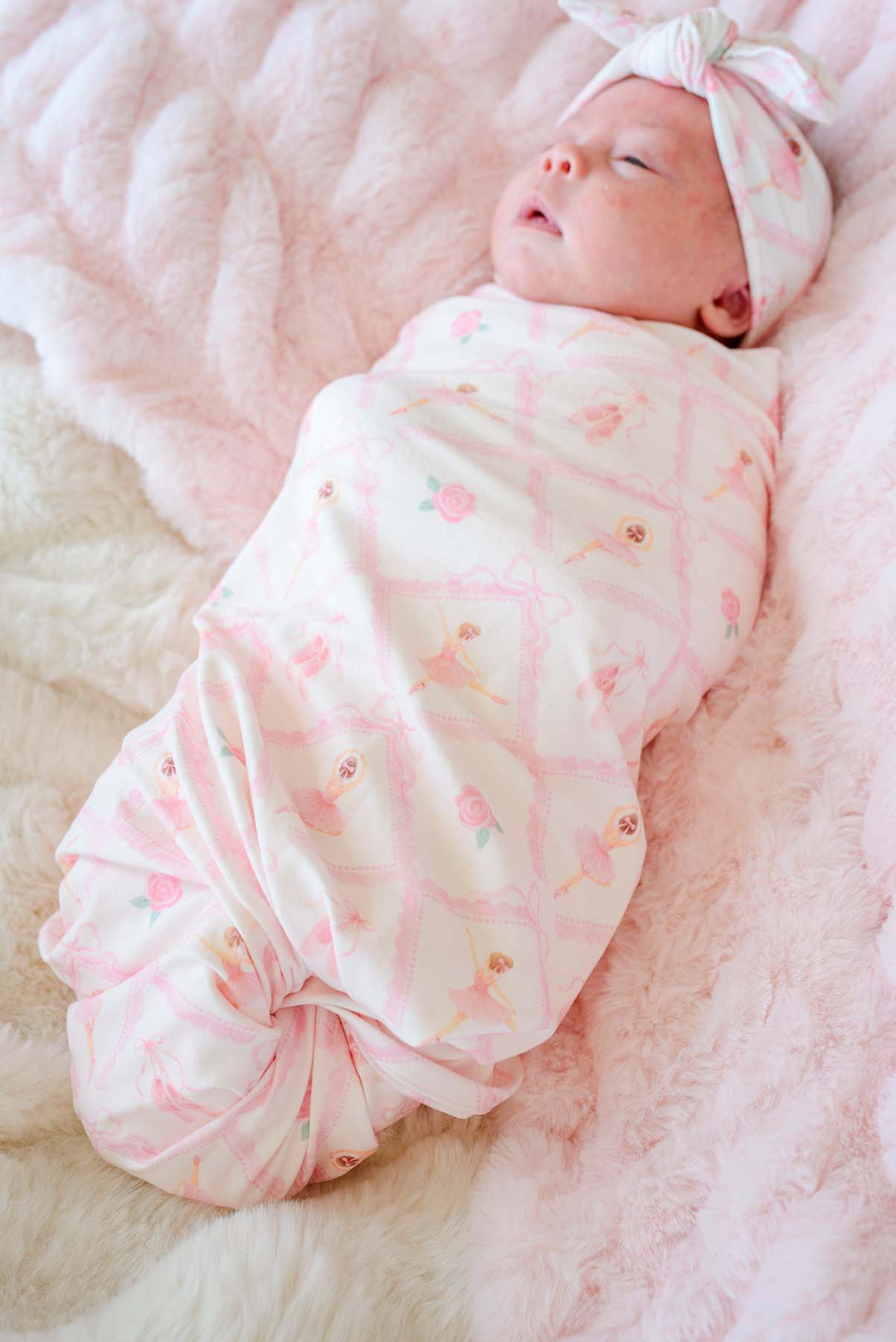 Sleeping newborn baby swaddled in a Caden Lane ballerina and roses pink print swaddle blanket with matching bow headwrap, laying next to a stuffed bunny on a pink fur blanket