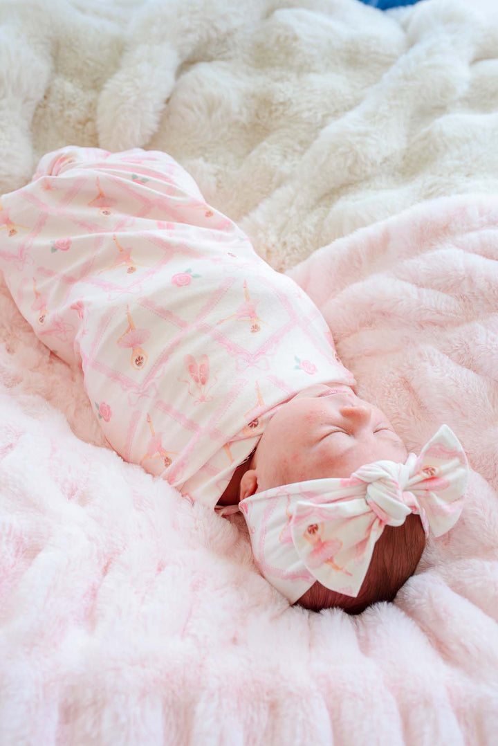 Close-up of sleeping newborn baby wearing a Caden Lane ballerina and roses pink print swaddle blanket with matching bow headwrap, laying on a pink faux fur blanket