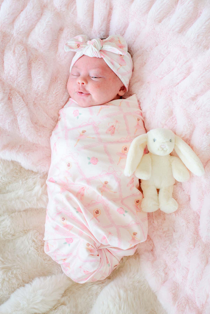 Sleeping newborn baby swaddled in a Caden Lane ballerina and roses pink print swaddle blanket with matching bow headwrap, laying next to a stuffed bunny on a pink fur blanket