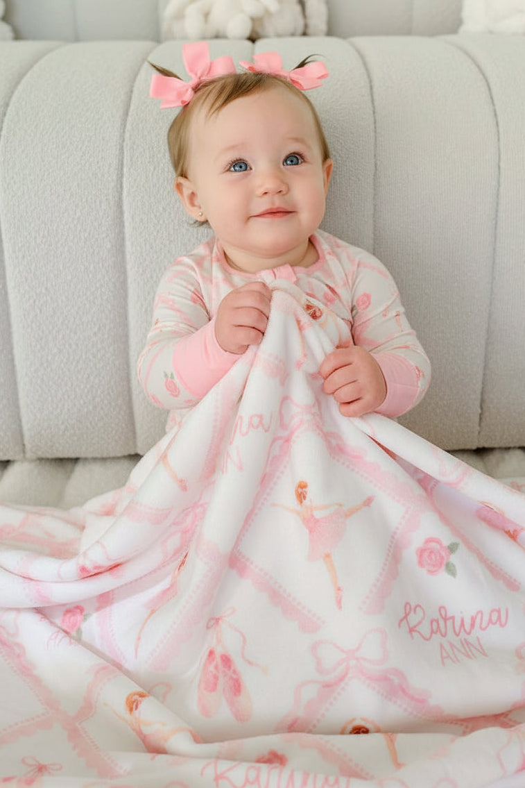 Baby girl in Caden Lane ballerina and roses pink print pajamas sitting with a matching personalized plush blanket embroidered with her name