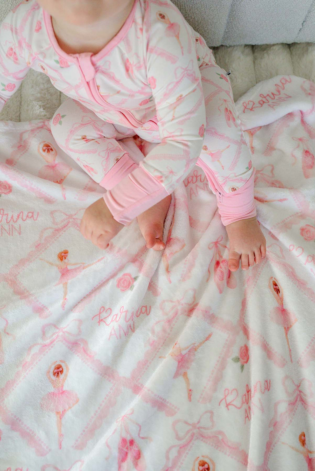 Baby in Caden Lane ballerina and roses pink print pajamas sitting with a matching personalized plush blanket embroidered with her name