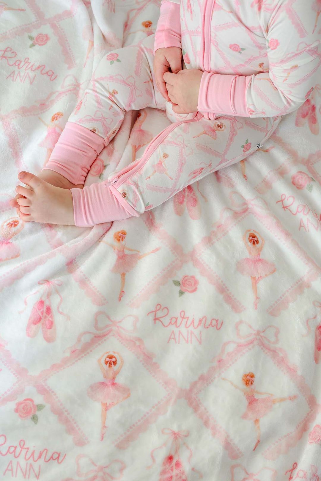 Baby in Caden Lane ballerina and roses pink print pajamas sitting with a matching personalized plush blanket embroidered with her name