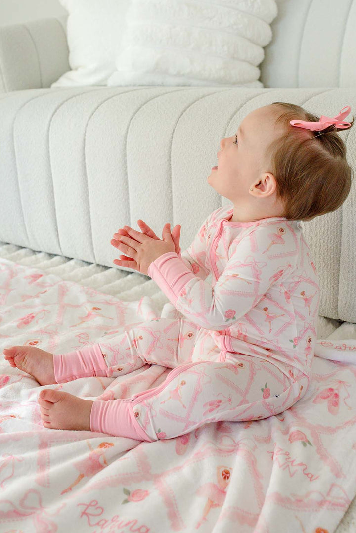 Baby girl in Caden Lane ballerina and roses pink print pajamas sitting with a matching personalized plush blanket embroidered with her name