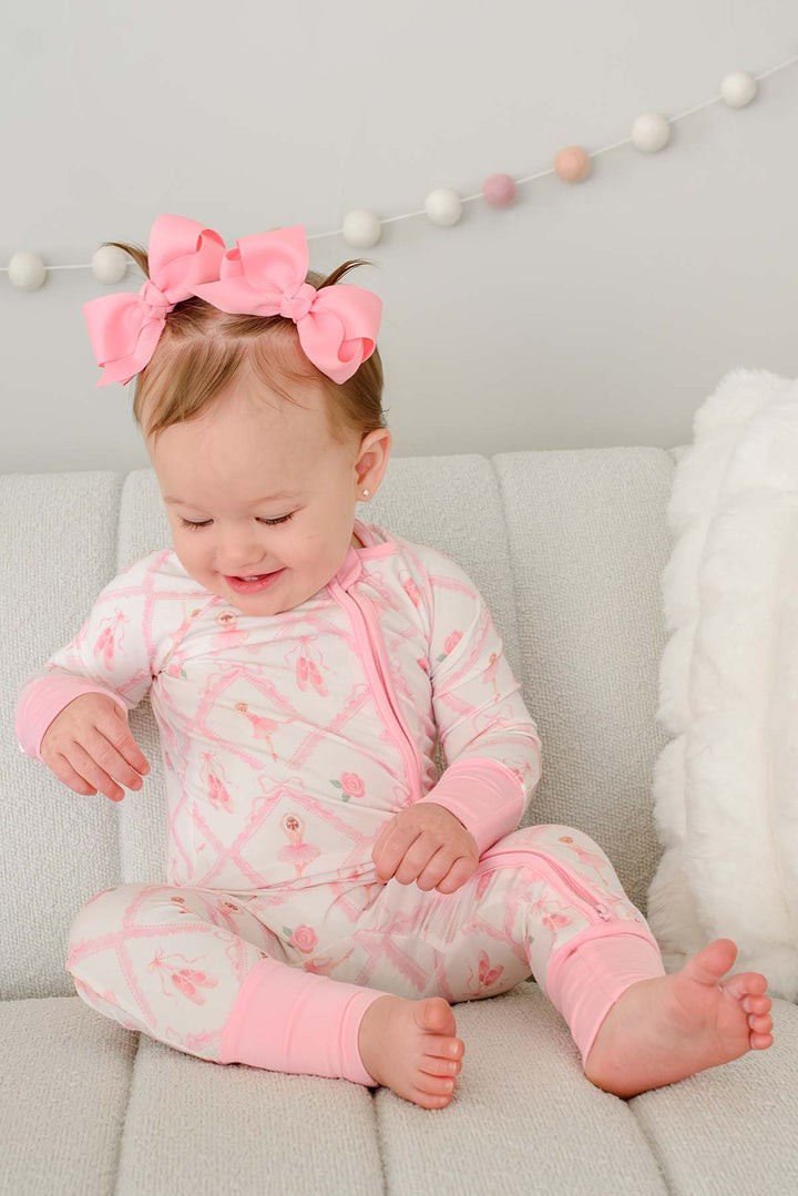 Toddler girl with pigtail pink bows sitting wearing a Caden Lane ballerina and roses pink print zip-up romper with pink cuffs