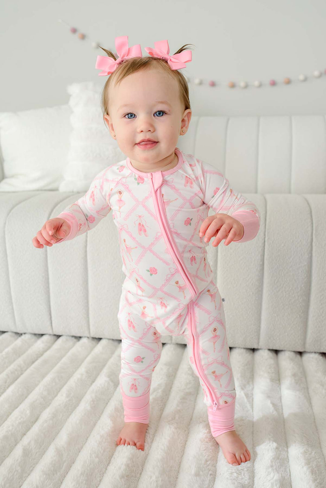 Toddler girl with pigtail pink bows standing wearing a Caden Lane ballerina and roses pink print zip-up romper with pink cuffs