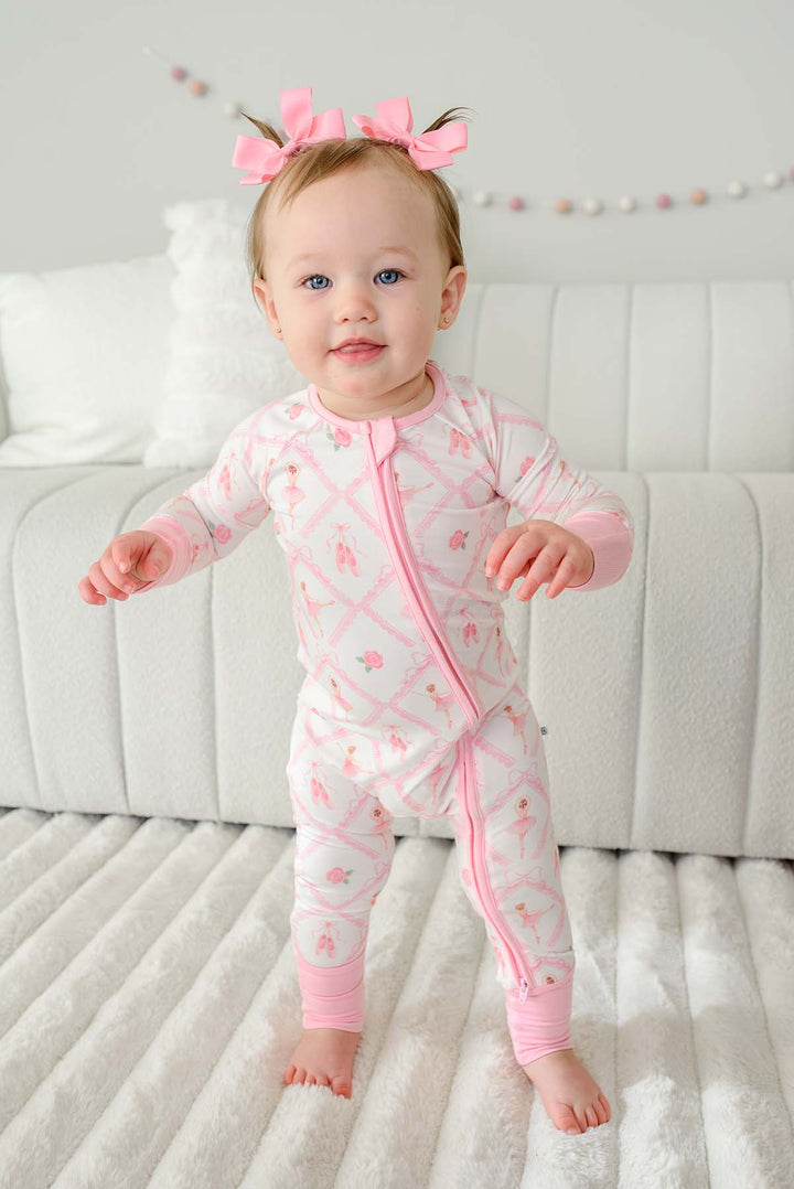 Toddler girl with pigtail pink bows standing wearing a Caden Lane ballerina and roses pink print zip-up romper with pink cuffs