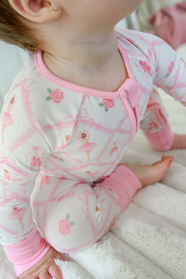 Toddler girl sitting wearing a Caden Lane ballerina and roses pink print zip-up romper with pink cuffs