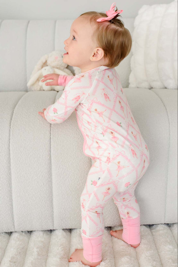Toddler girl with pigtail pink bows standing wearing a Caden Lane ballerina and roses pink print zip-up romper with pink cuffs