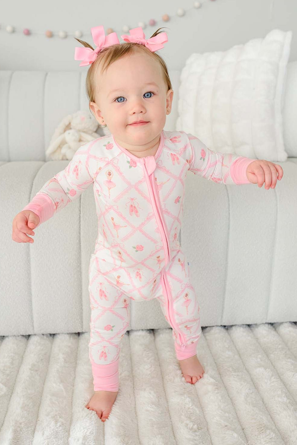 Toddler girl with pigtail pink bows standing wearing a Caden Lane ballerina and roses pink print zip-up romper with pink cuffs