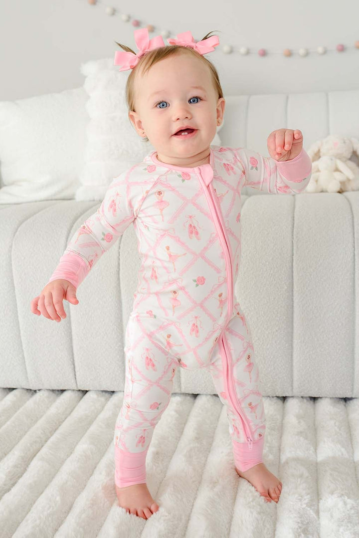 Toddler girl with pigtail pink bows standing wearing a Caden Lane ballerina and roses pink print zip-up romper with pink cuffs