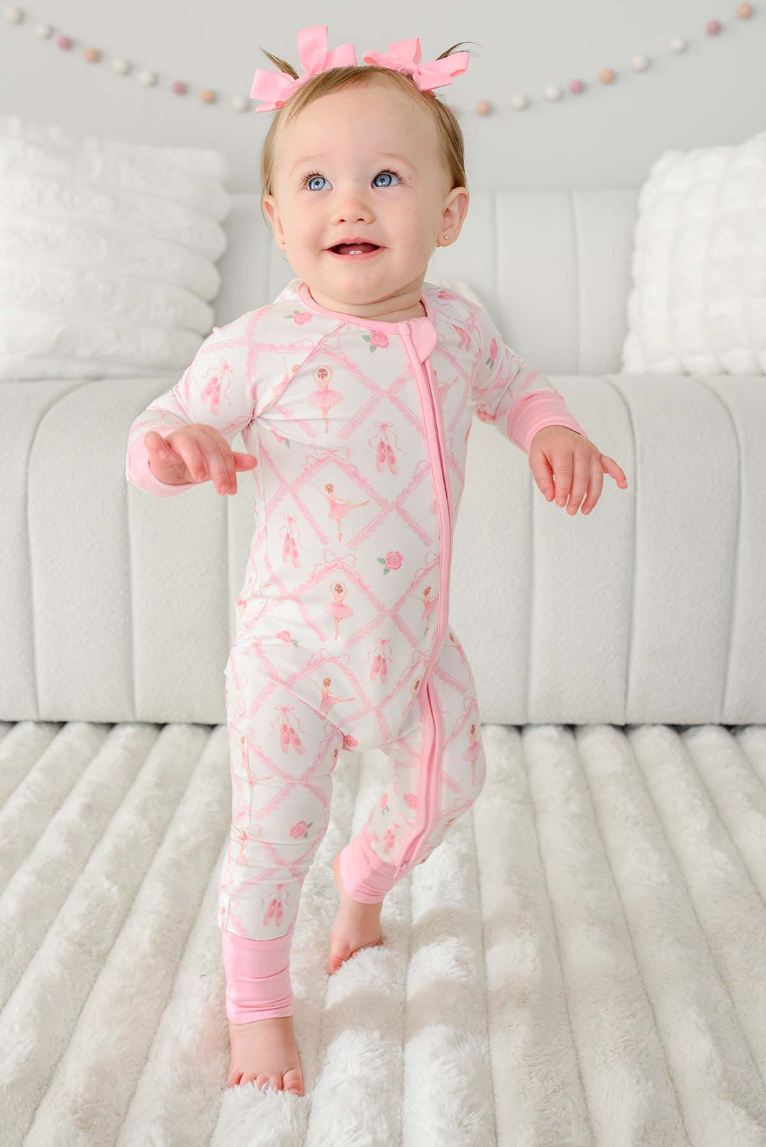 Toddler girl with pigtail pink bows standing wearing a Caden Lane ballerina and roses pink print zip-up romper with pink cuffs