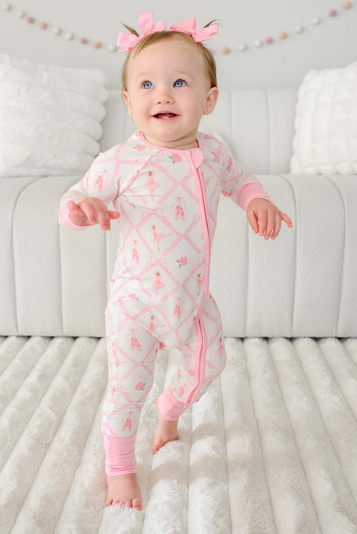 Toddler girl with pigtail pink bows standing wearing a Caden Lane ballerina and roses pink print zip-up romper with pink cuffs