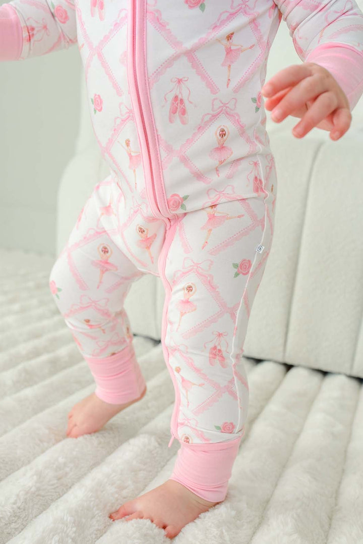 Toddler girl standing wearing a Caden Lane ballerina and roses pink print zip-up romper with pink cuffs