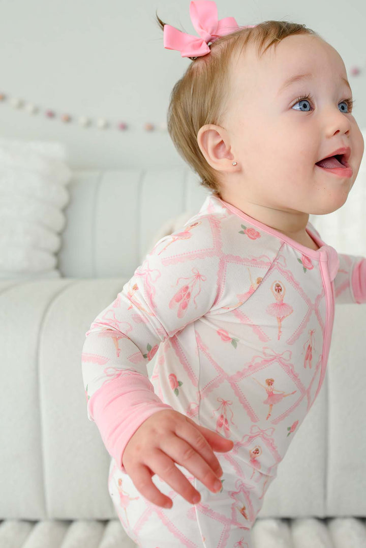 Toddler girl with pigtail pink bows standing wearing a Caden Lane ballerina and roses pink print zip-up romper with pink cuffs