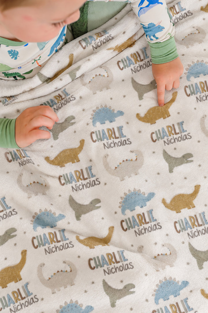Close-up of two toddlers in dinosaur-print pajamas pointing at a personalized Caden Lane Dino Stomps blanket, showing dinosaur silhouettes and custom name print