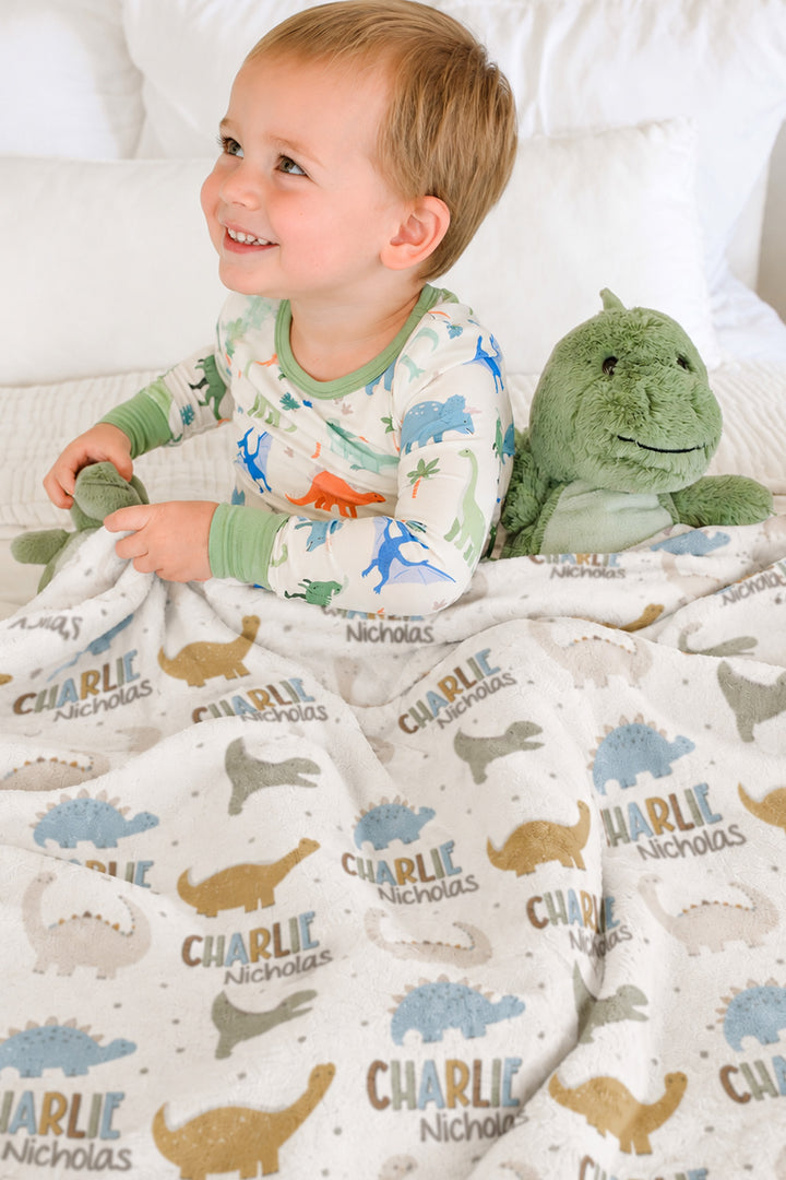 Smiling toddler in dinosaur-print pajamas sitting on a bed with a plush dinosaur toy, wrapped in a personalized Caden Lane Dino Stomps blanket
