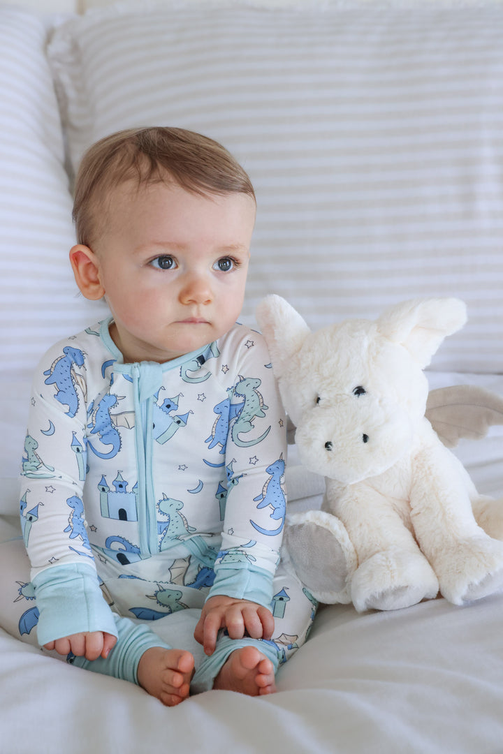 baby boy sitting on bed in dragon pajama romper with a stuffed white dragon stuffed animal 