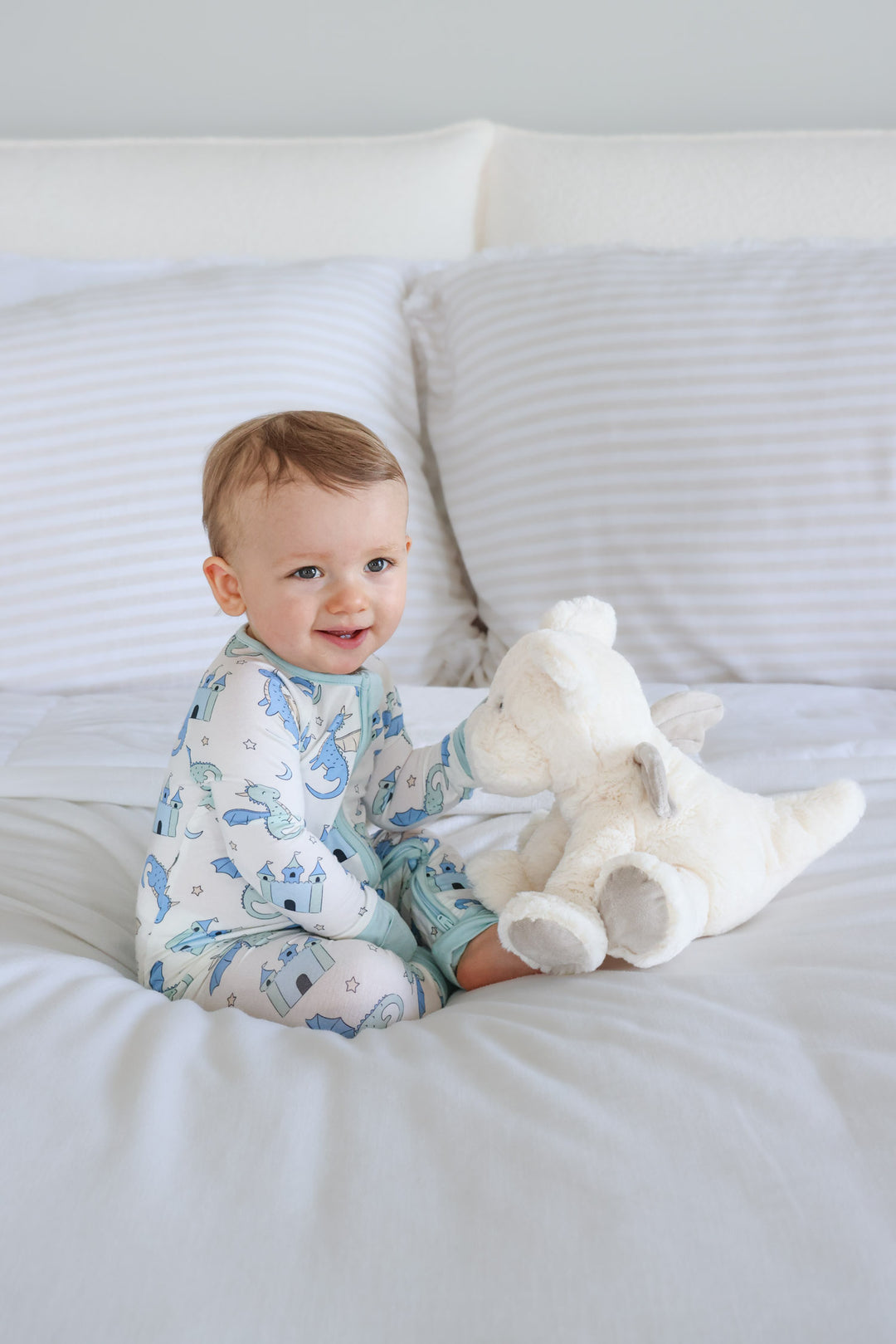 smiling baby boy sitting on bed with white stuffed dragon