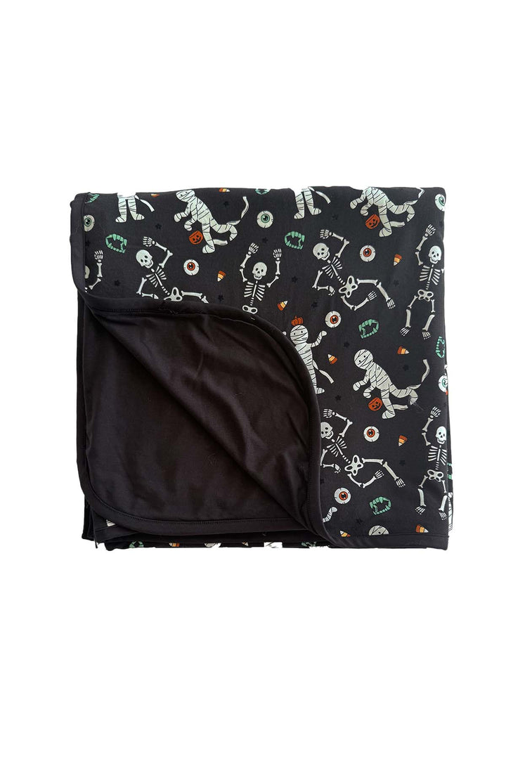 Caden Lane bamboo blanket in black, folded to show the solid black backing and a Halloween skeleton, mummy, and candy corn print, with vampire fangs and eyeball accents throughout