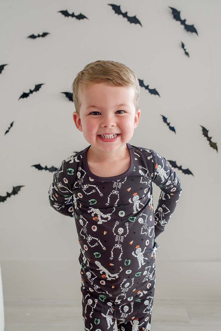 Boy smiling, standing and wearing a Caden Lane black two-piece pajama set with a white skeleton, mummy, and candy corn print, with Halloween bat decorations in the background