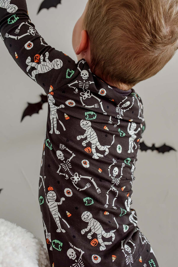 Boy standing and wearing a Caden Lane black two-piece pajama set with a white skeleton, mummy, and candy corn print, with Halloween bat decorations in the background