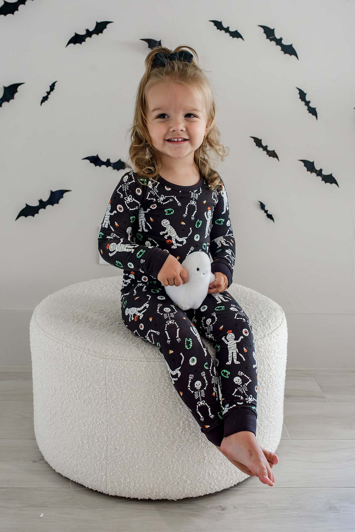 Girl holding a plush ghost and wearing a Caden Lane black two-piece pajama set with a white skeleton, mummy, and candy corn print, with Halloween bat decorations in the background