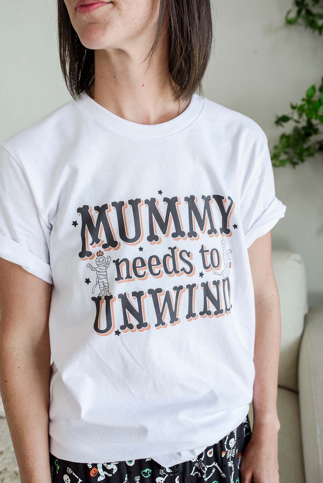 A woman wearing a white short-sleeve graphic T-shirt with the text 'MUMMY needs to UNWIND' in dark lettering with a peach outline, paired with black patterned shorts.