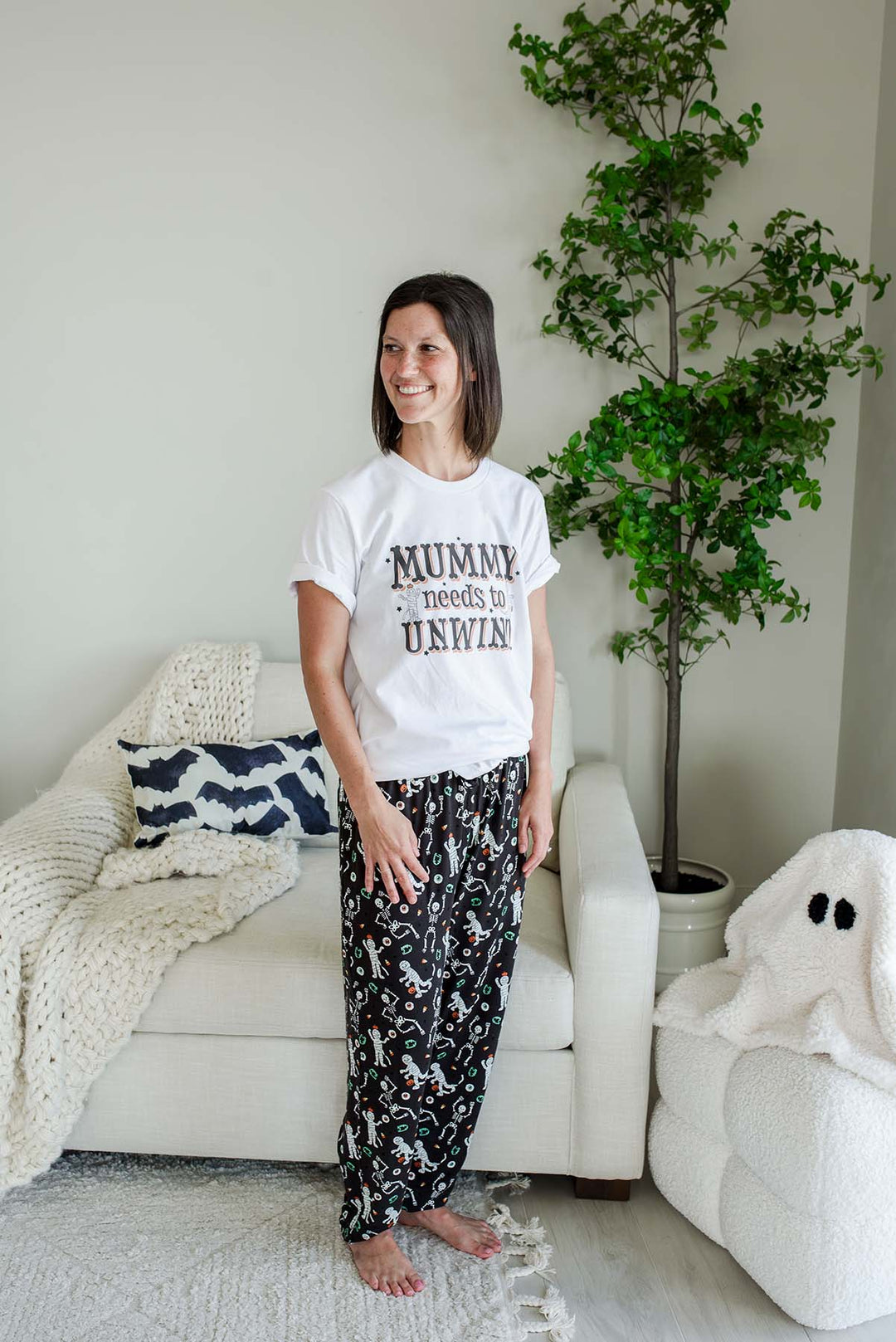 A woman stands in a living room wearing a white graphic T-shirt that reads 'MUMMY needs to UNWIND' and black patterned pajama pants. The room has a white sofa with a knit blanket, a potted tree, and a white plush ghost on the floor.