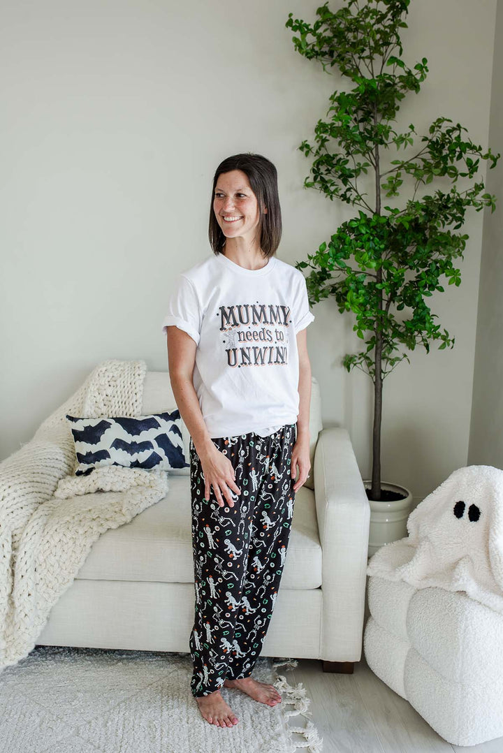 A woman stands in a living room wearing a white graphic T-shirt that reads 'MUMMY needs to UNWIND' and black patterned pajama pants. The room has a white sofa with a knit blanket, a potted tree, and a white plush ghost on the floor.