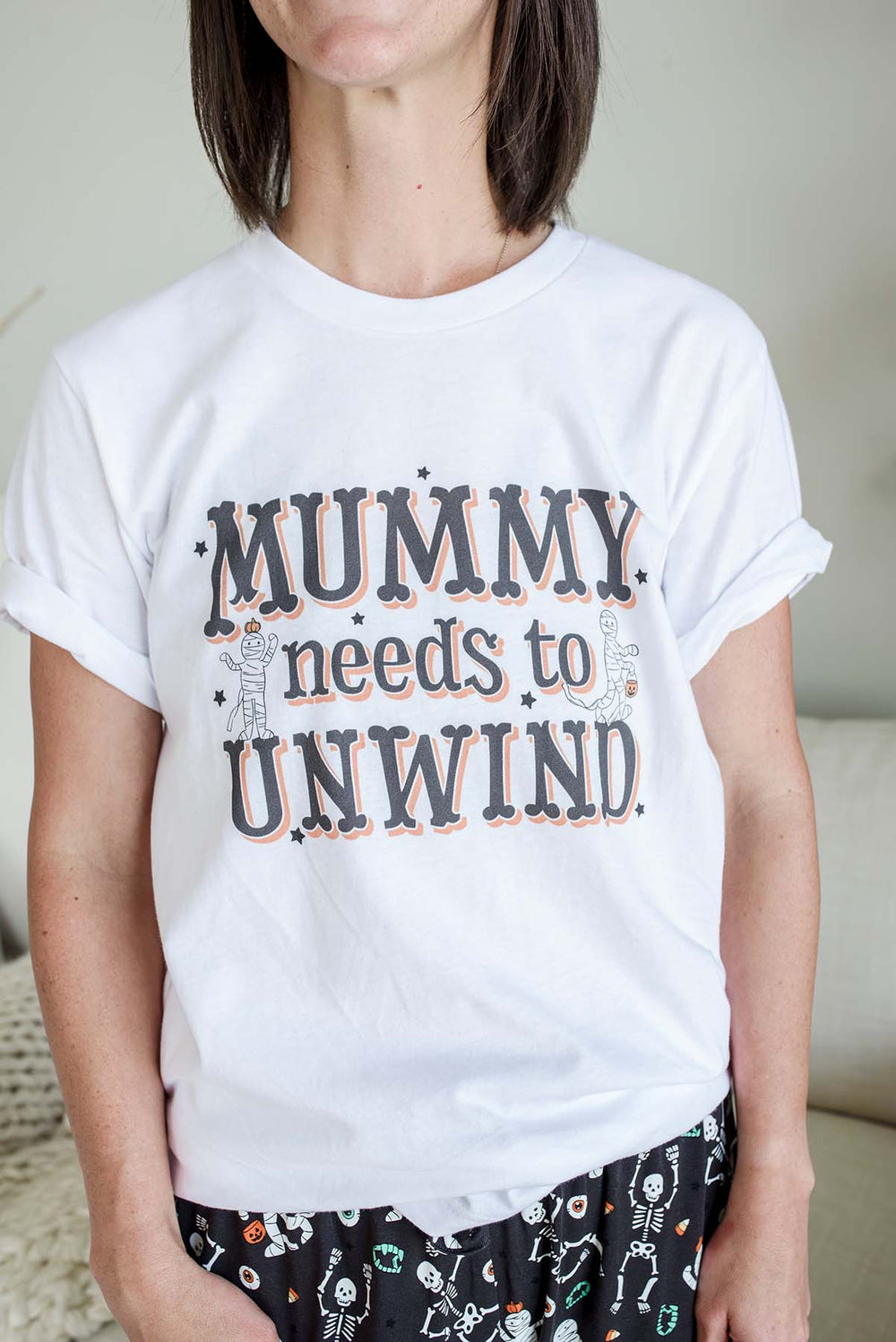 Mom wearing White Caden Lane Halloween t-shirt for women featuring 'Mummy Needs to Unwind' text in black and orange lettering with two cartoon mummy characters holding pumpkin treat buckets, surrounded by black star accents