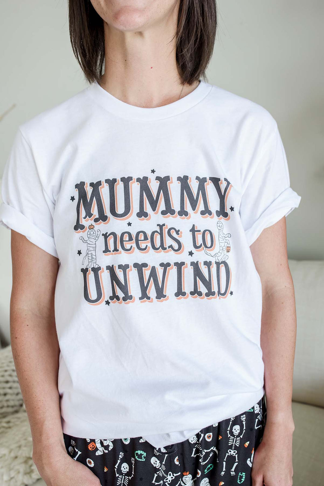 Mom wearing White Caden Lane Halloween t-shirt for women featuring 'Mummy Needs to Unwind' text in black and orange lettering with two cartoon mummy characters holding pumpkin treat buckets, surrounded by black star accents