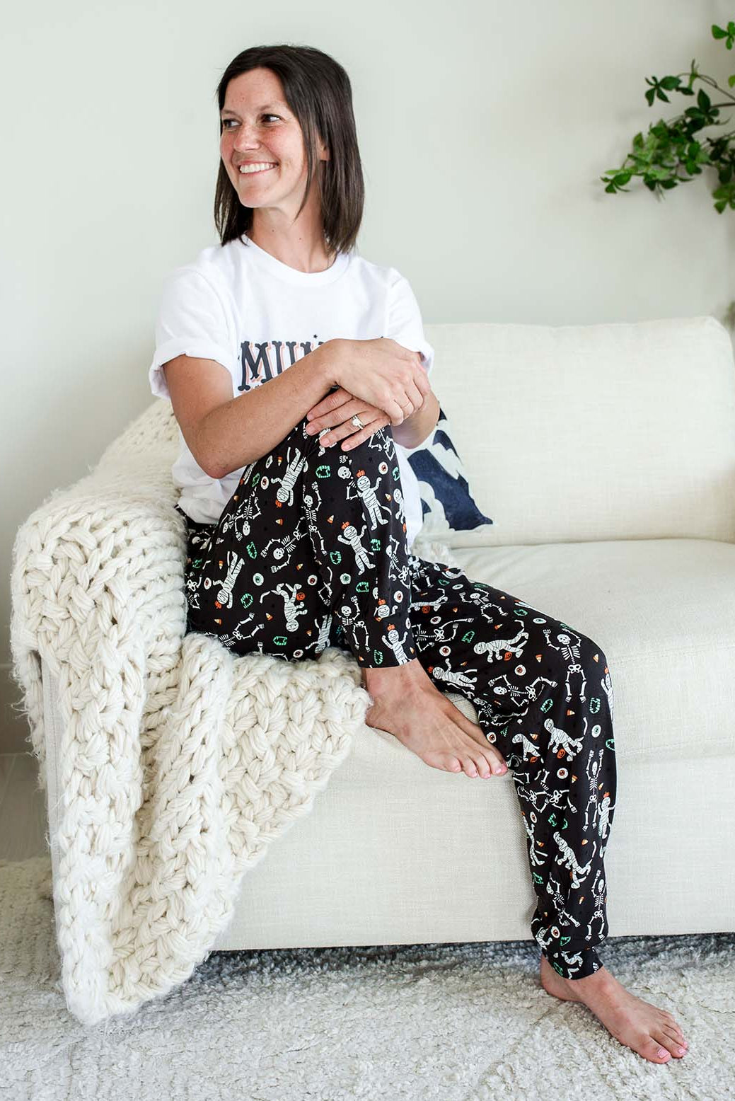 Adult wearing Caden Lane black Halloween pajama pants with white skeleton, mummy, and candy corn print, paired with a white 'Mummy needs to Unwind' graphic tee