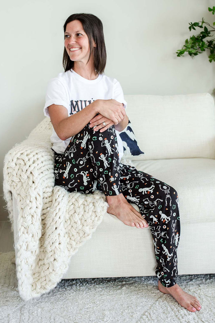 Adult wearing Caden Lane black Halloween pajama pants with white skeleton, mummy, and candy corn print, paired with a white 'Mummy needs to Unwind' graphic tee