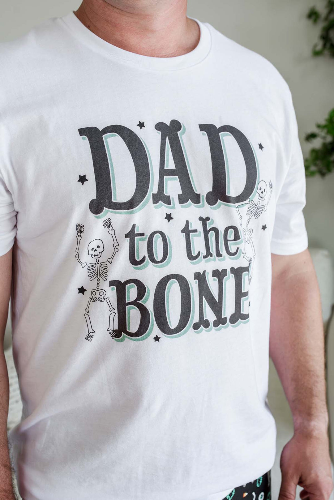 A man wearing a white crewneck t-shirt with a graphic that reads 'DAD to the BONE' in dark gray letters with a light green shadow, decorated with cartoon skeletons and small stars.