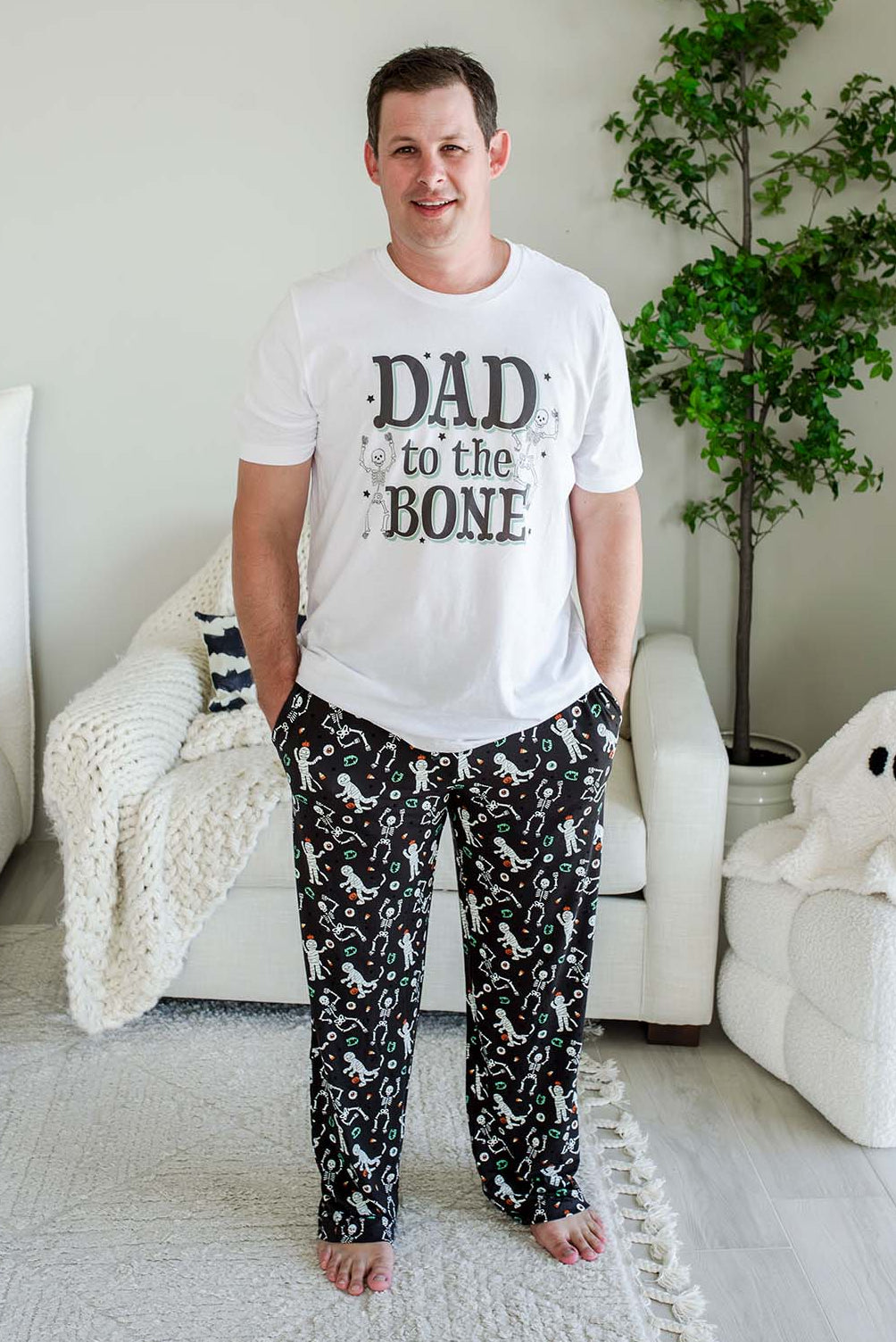 A man standing in a bedroom wearing a white graphic t-shirt that reads 'DAD to the BONE' and black pajama pants with a white skeleton print.