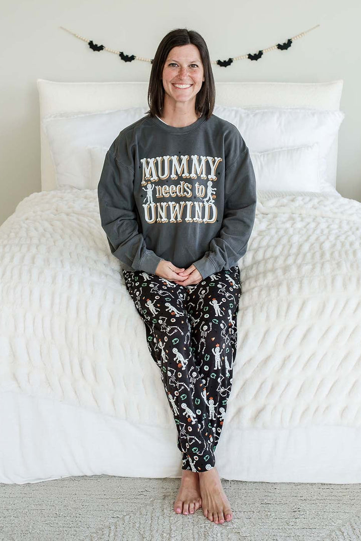 Adult wearing Caden Lane black Halloween pajama pants with white skeleton, mummy, and candy corn print, paired with a white 'Mummy needs to Unwind' graphic sweater
