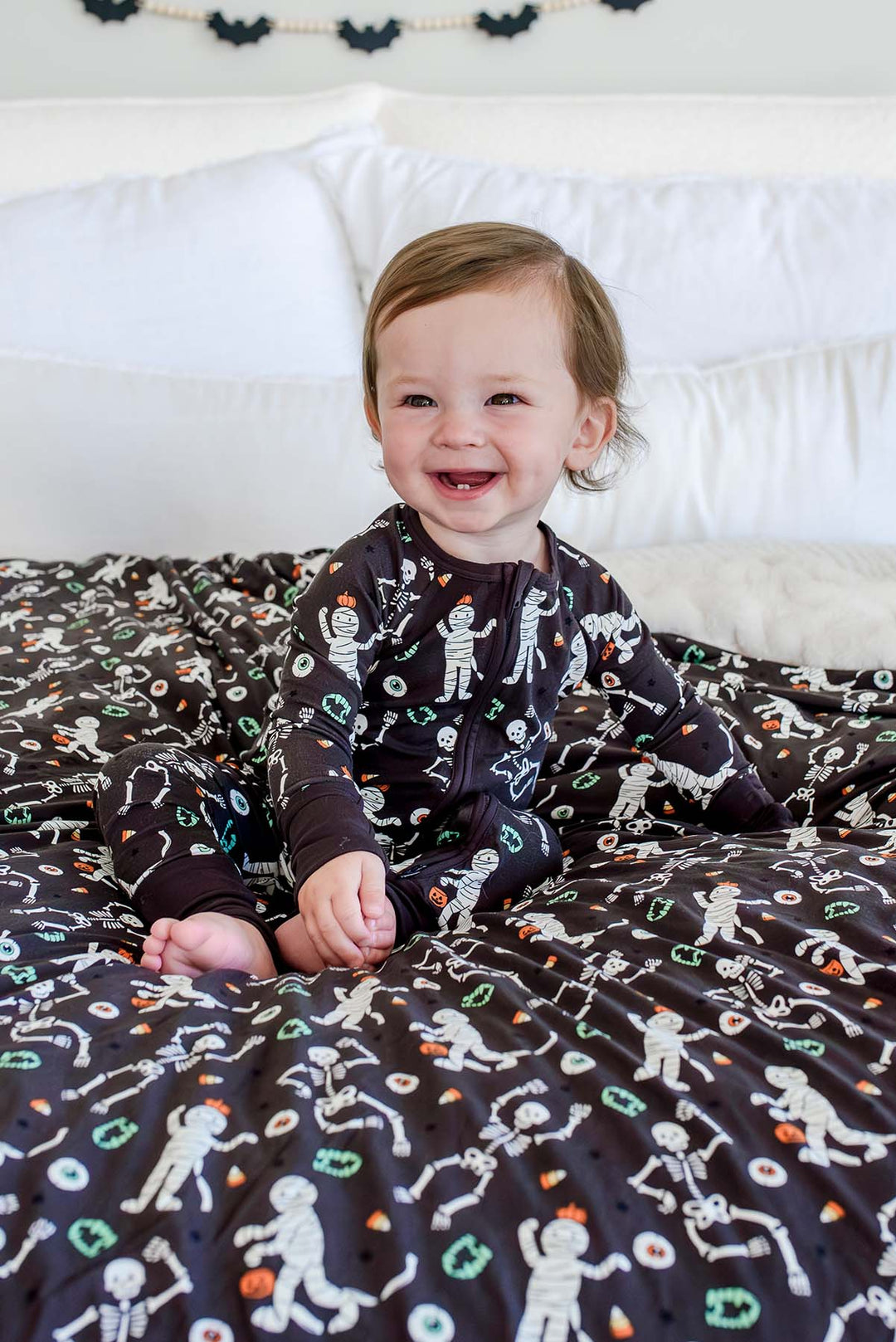 Smiling baby wearing a Caden Lane black zip-up skeleton and mummy print pajama sitting on a matching black Caden Lane bamboo blanket with the same Halloween print.