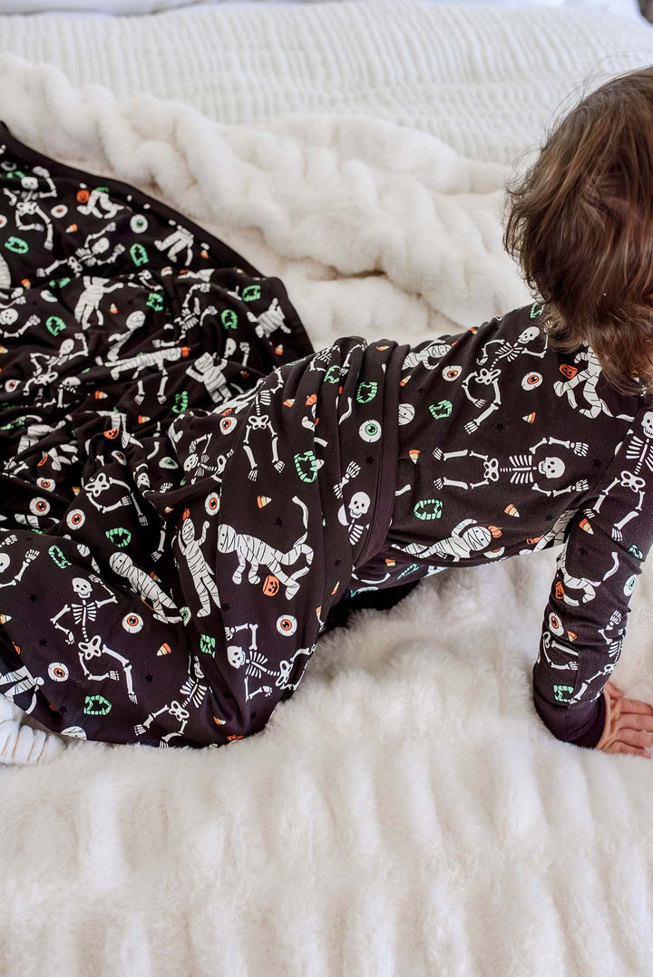 Baby wearing a Caden Lane black zip-up skeleton and mummy print pajama sitting on a matching black Caden Lane bamboo blanket with the same Halloween print. On a white bed with a Caden Lane winter white faux fur blanket.