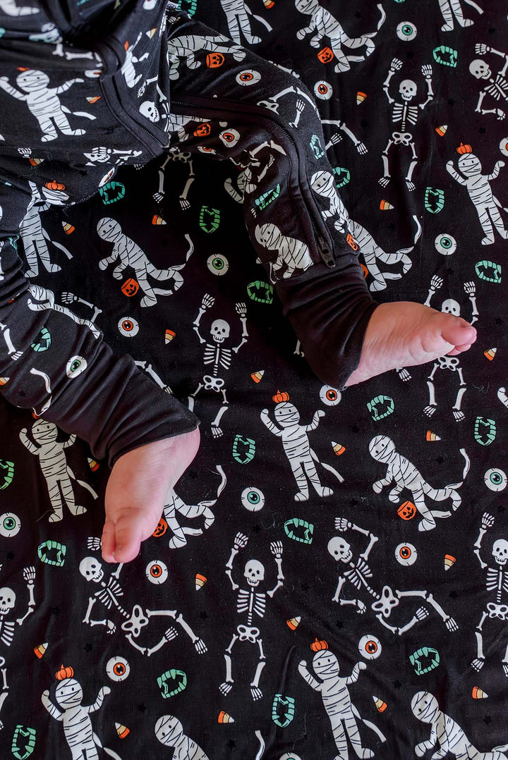 Baby wearing a Caden Lane black zip-up skeleton and mummy print pajama sitting on a matching black Caden Lane bamboo blanket with the same Halloween print.