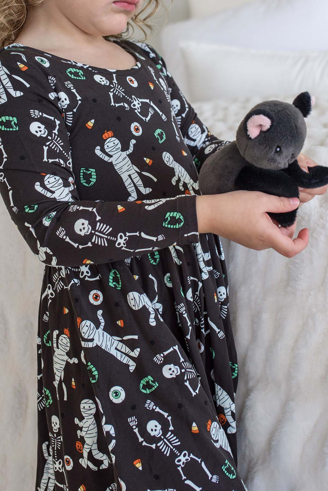 Young girl holding a stuffed bat and wearing a Caden Lane black long-sleeve Halloween dress with a twirl skirt, featuring a white skeleton, mummy, and candy corn print