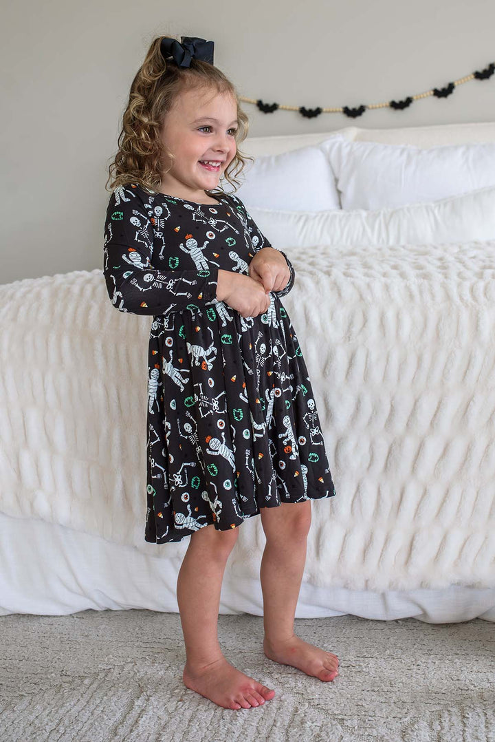 Young girl wearing a Caden Lane black long-sleeve Halloween dress with a twirl skirt, featuring a white skeleton, mummy, and candy corn print, styled with a black hair bow. White bed with bat garland behind her.