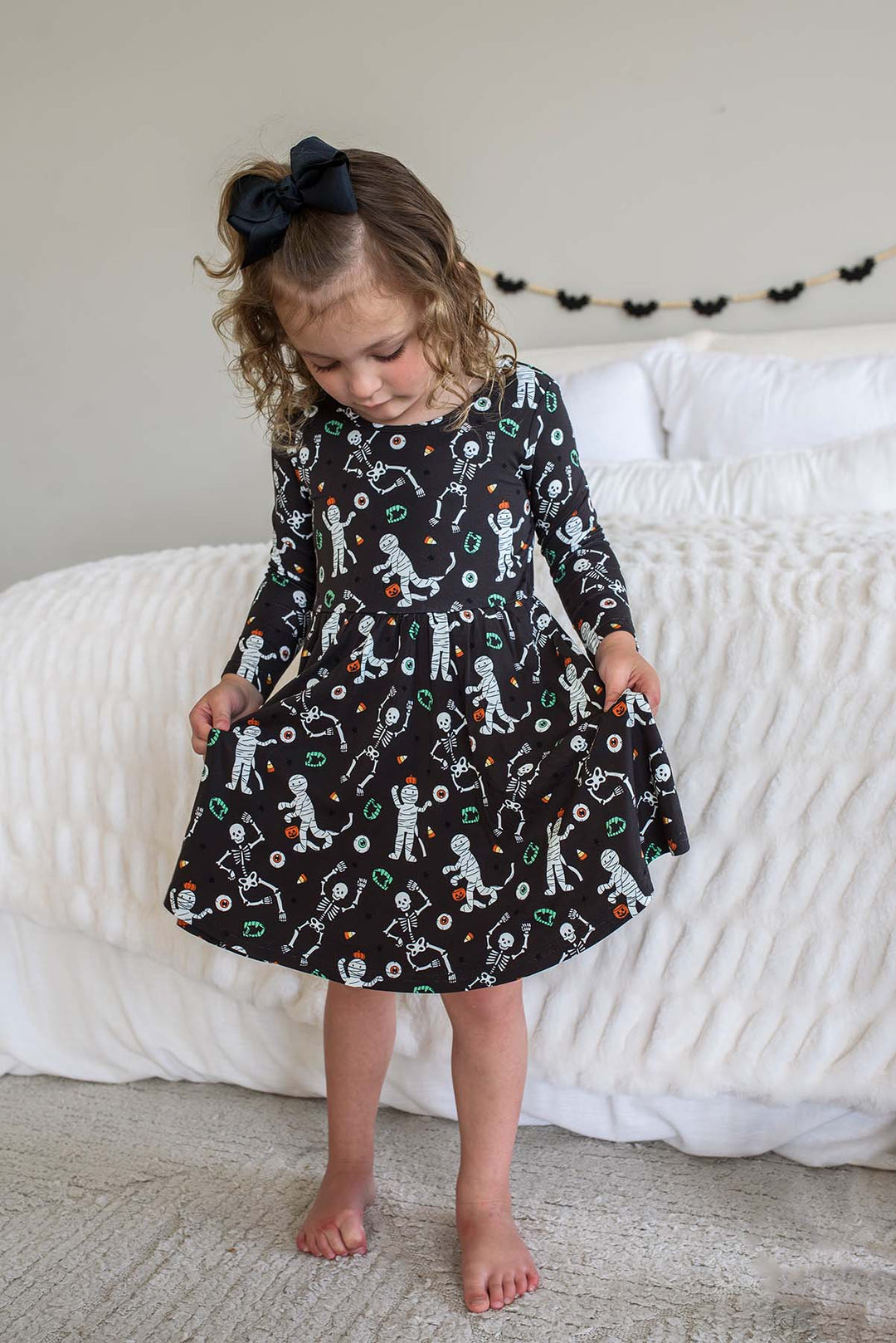 Young girl wearing a Caden Lane black long-sleeve Halloween dress with a twirl skirt, featuring a white skeleton, mummy, and candy corn print, styled with a black hair bow. White bed with bat garland behind her.