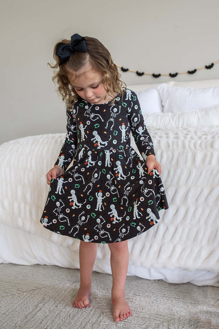 Young girl wearing a Caden Lane black long-sleeve Halloween dress with a twirl skirt, featuring a white skeleton, mummy, and candy corn print, styled with a black hair bow. White bed with bat garland behind her.