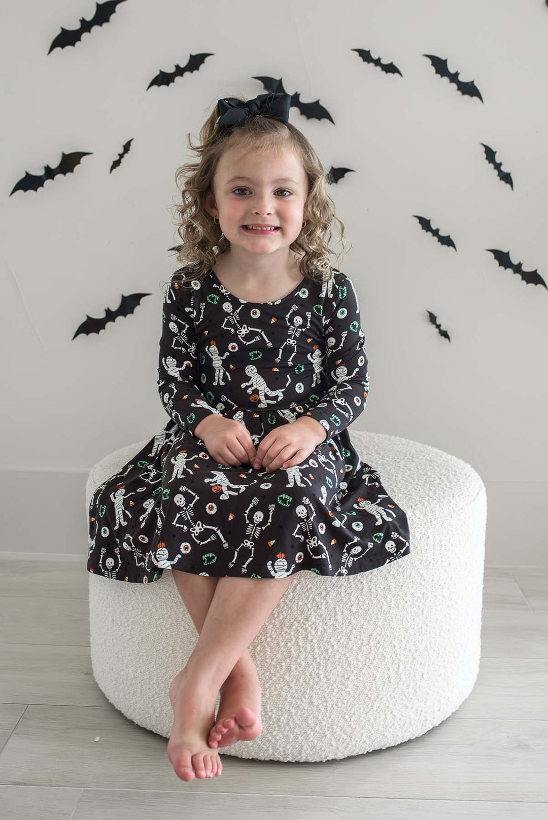 Young girl wearing a Caden Lane black long-sleeve Halloween dress with a twirl skirt, featuring a white skeleton, mummy, and candy corn print, styled with a black hair bow. With bat cutouts on white wall behind her.