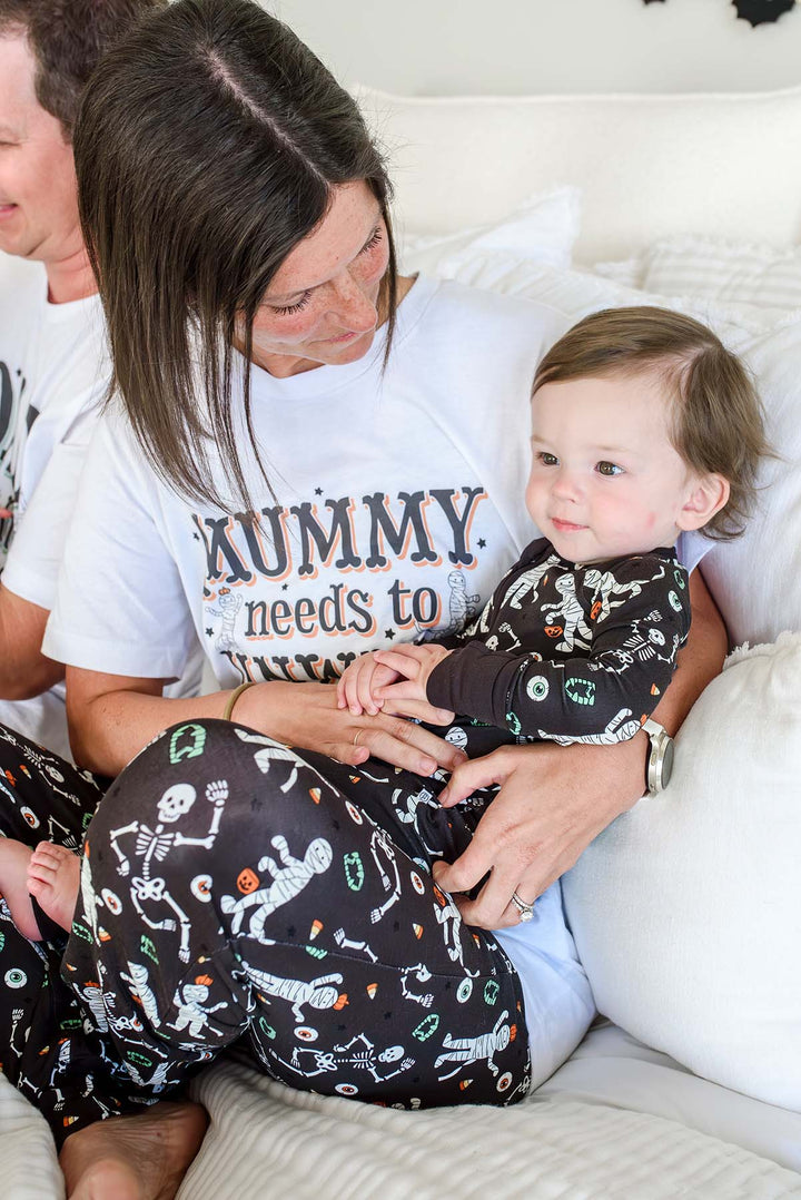 A woman and a toddler wearing matching skeleton-print pajamas and a white graphic T-shirt while sitting on a couch.