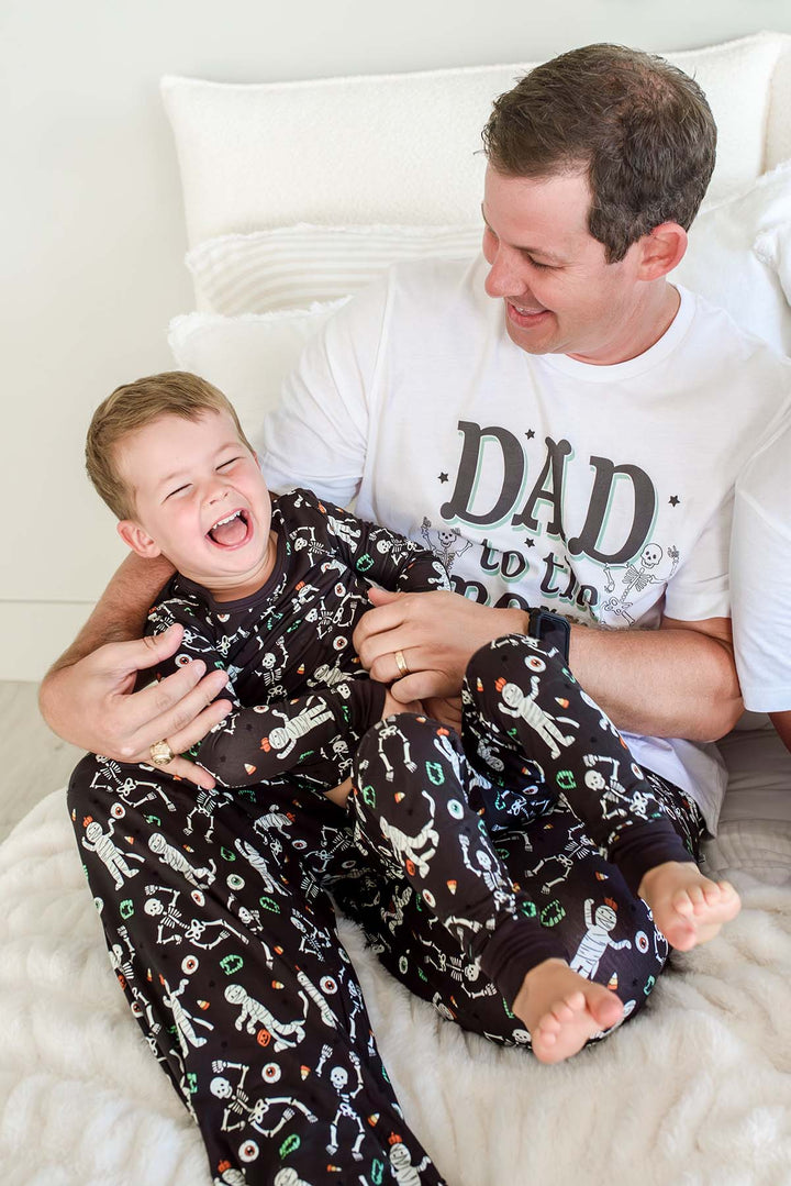 Father and son laughing together on a bed, both wearing matching 'Dad to the Bone' Halloween pajamas with a white skeleton print on black fabric.