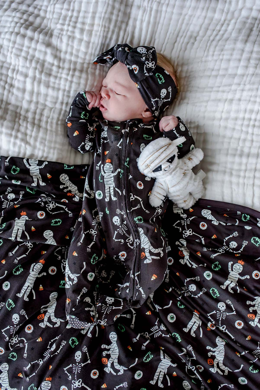 Sleeping newborn wearing a Caden Lane black skeleton and mummy print footie with matching bow headband, lying on the coordinating blanket and holding a plush mummy toy.