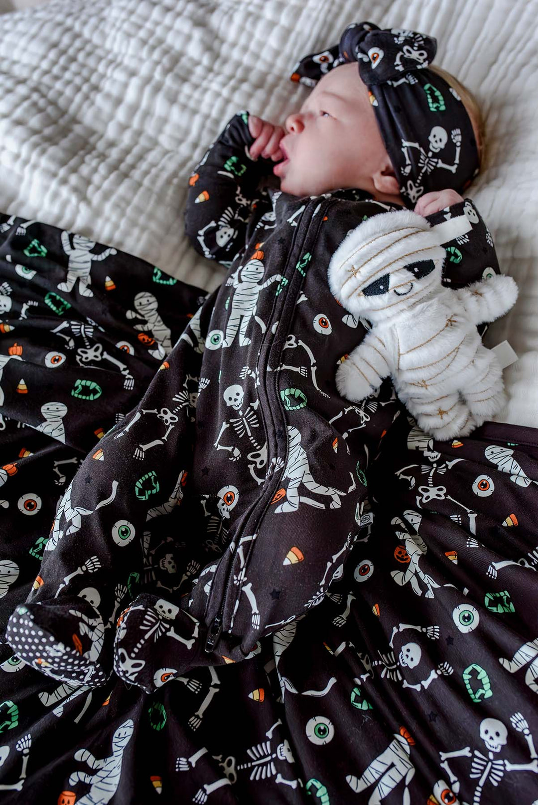 Sleeping newborn wearing a Caden Lane black skeleton and mummy print footie with matching bow headband, lying on the coordinating blanket and holding a plush mummy toy.