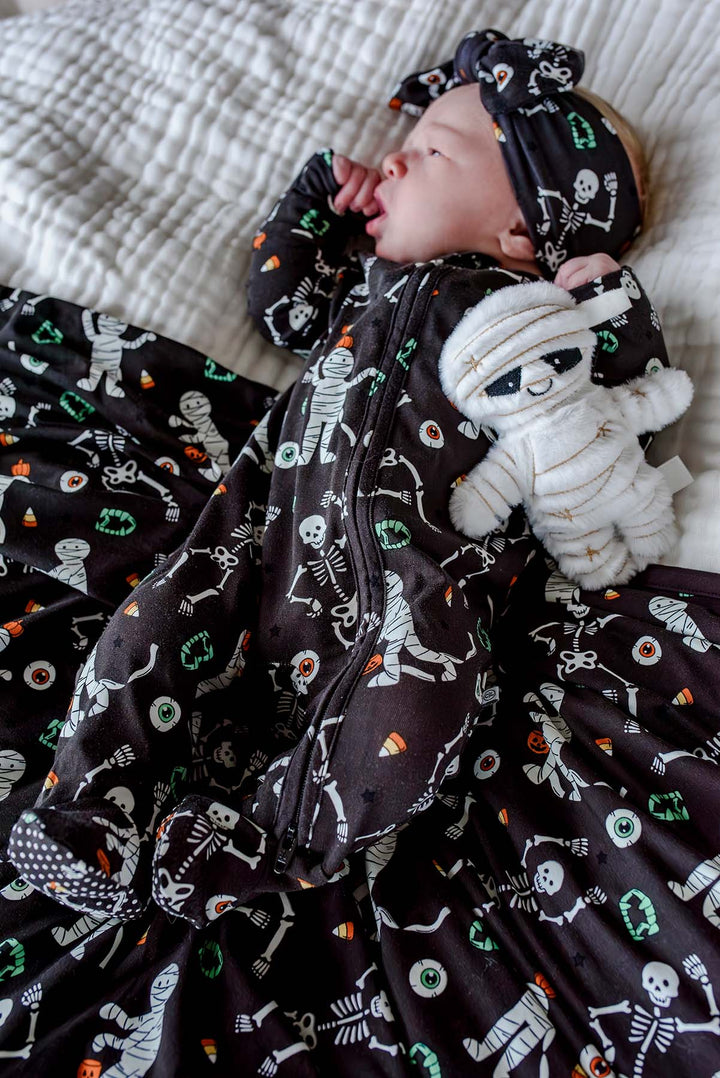 Sleeping newborn wearing a Caden Lane black skeleton and mummy print footie with matching bow headband, lying on the coordinating blanket and holding a plush mummy toy.